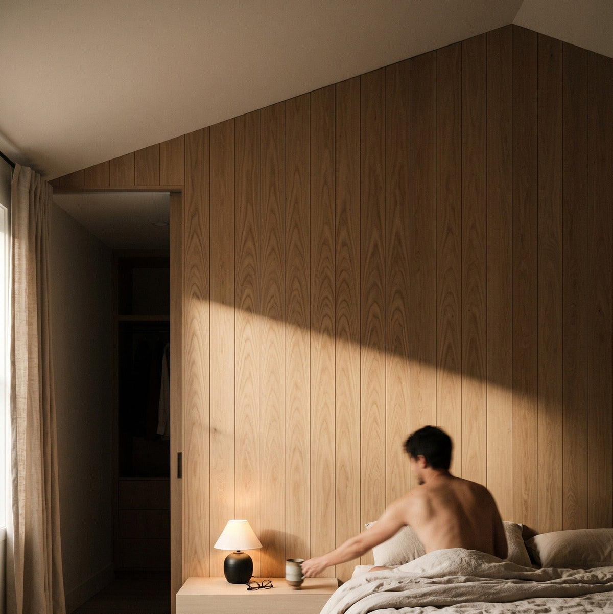 A man sits on a bed in a minimalist bedroom with light wood paneling, sunlight streaming across the wall, and a bedside table with a lit lamp.