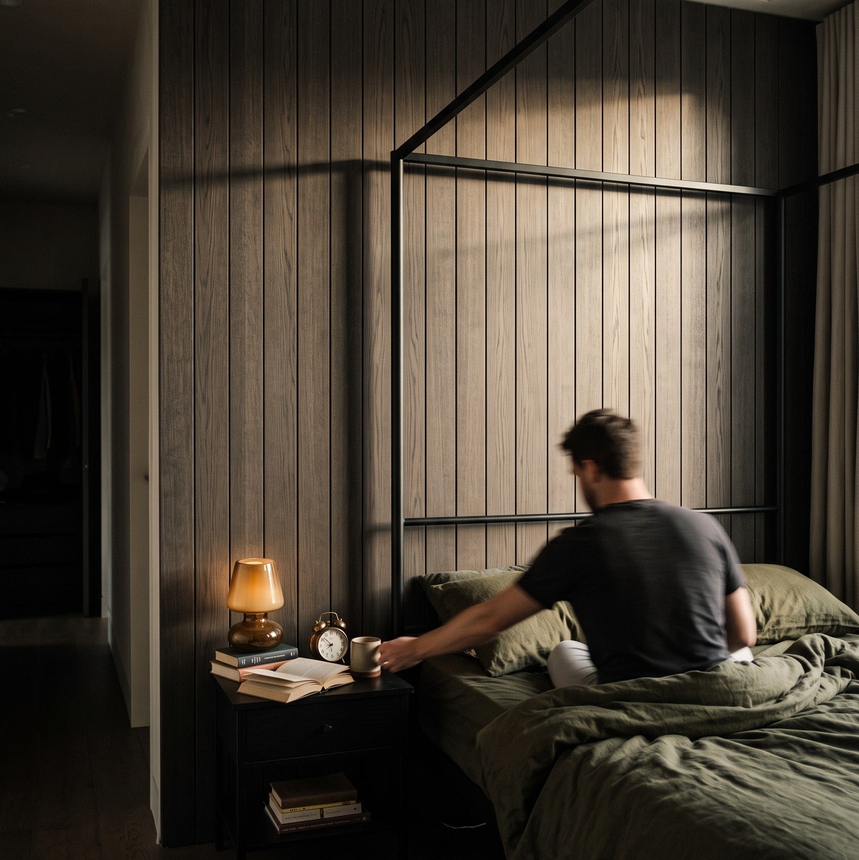 A man sits on a bed in a dark, modern bedroom with wood-paneled walls, a black metal canopy bed frame, and a bedside table with a lit lamp.