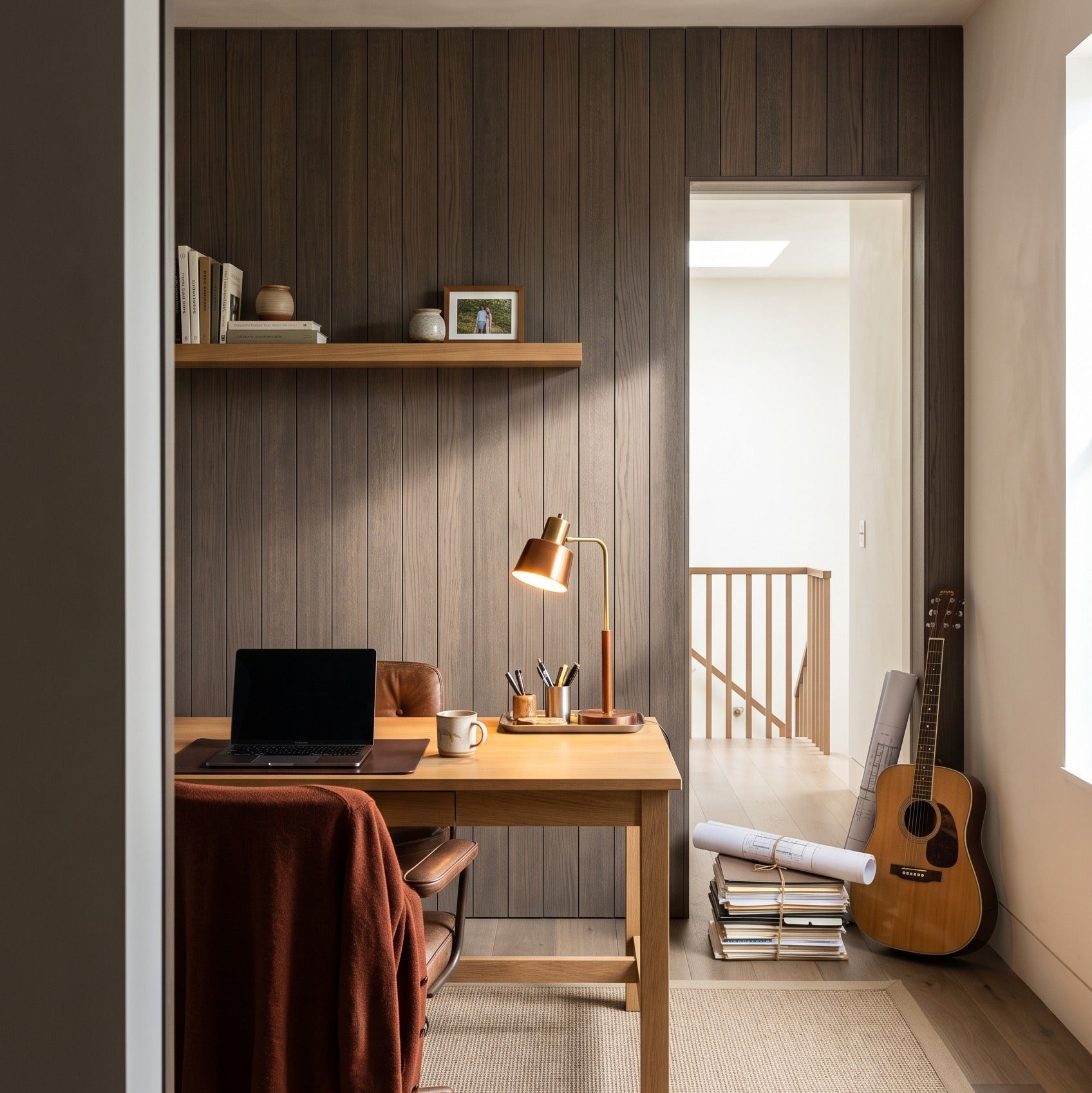 A modern home office interior featuring a light wood desk against a dark wood-paneled wall, with a laptop, lamp, and chair. An acoustic guitar rests on a stack of books near an open doorway, and a woven rug covers the floor.