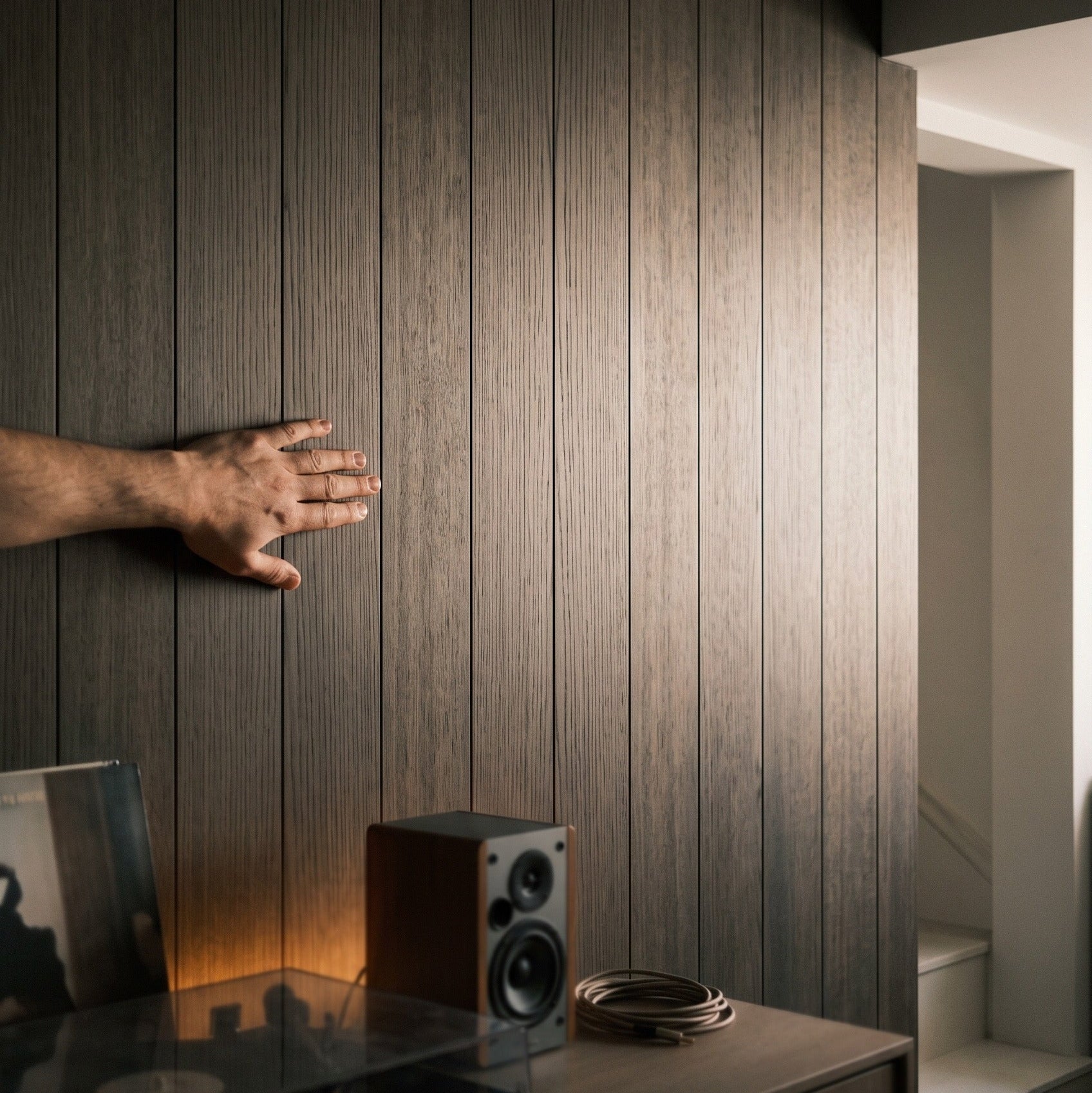 Dark brown wood panel wall with vertical planks, shown in a modern interior setting with a hand touching the surface, a record player, and a speaker on a console.