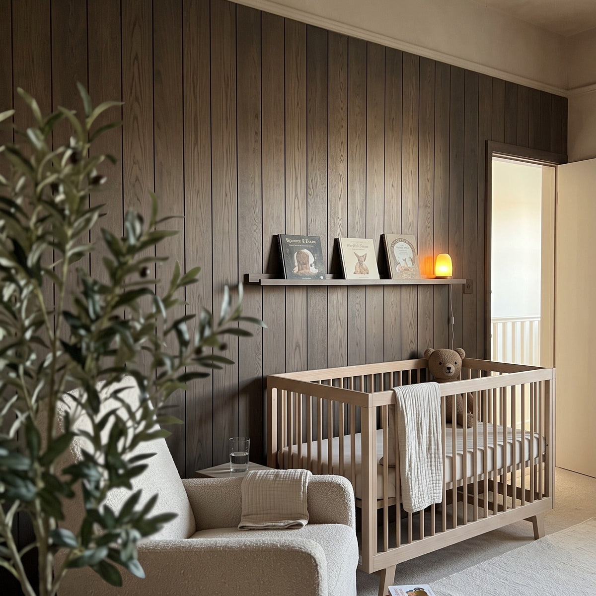 A styled nursery interior featuring a light wood crib, a beige upholstered armchair, a potted olive tree, and a dark wood-paneled wall with a floating shelf.