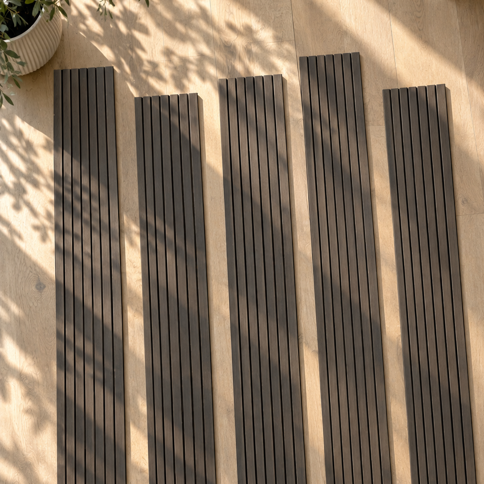 Five long dark gray textured slats, likely outdoor deck or patio boards, arranged vertically on a light wood floor with dappled sunlight and leaf shadows.