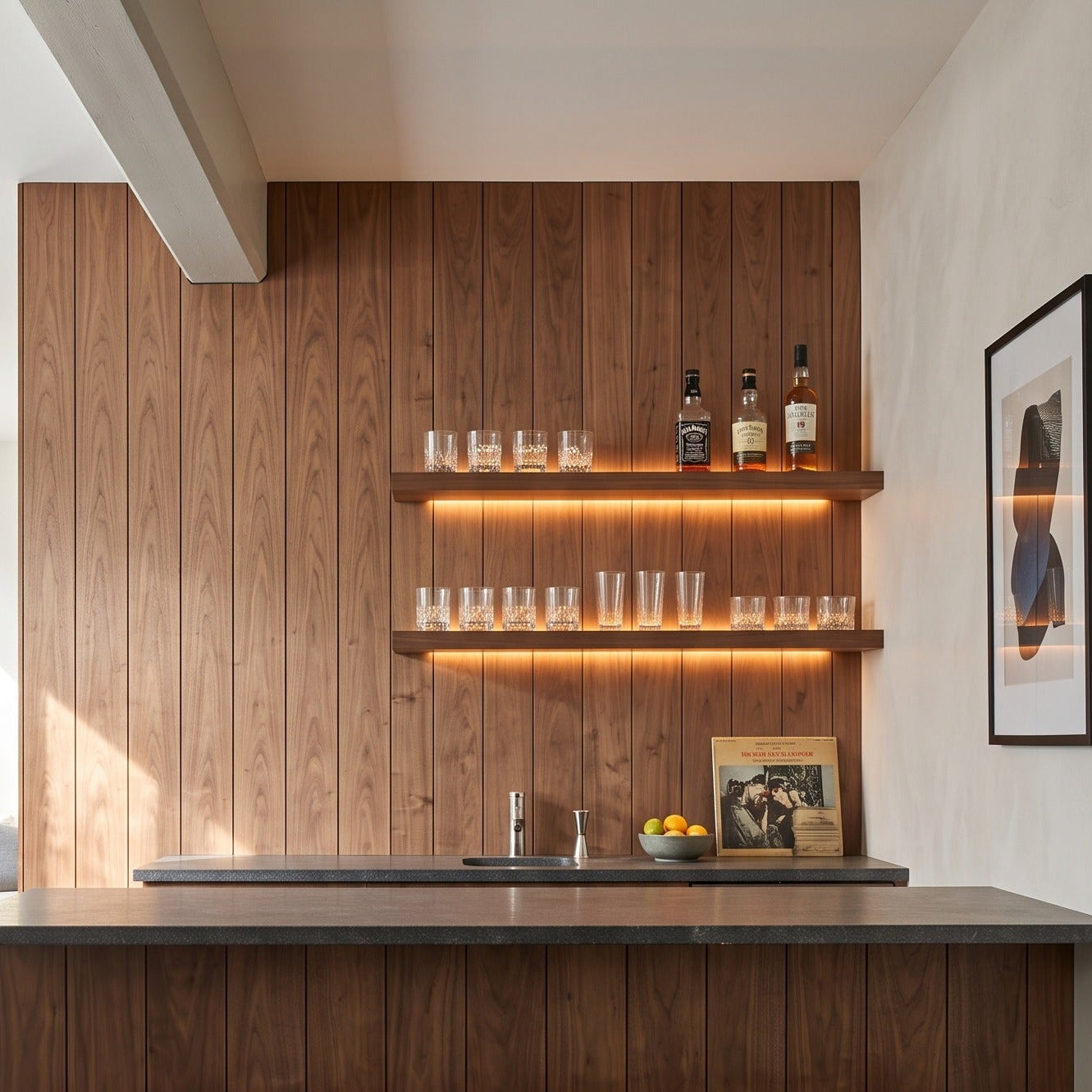 A modern kitchen island with a dark wood grain cabinet and countertop, three wooden bar stools, built-in open shelves with warm lighting, and a wood-paneled back wall in a bright interior.