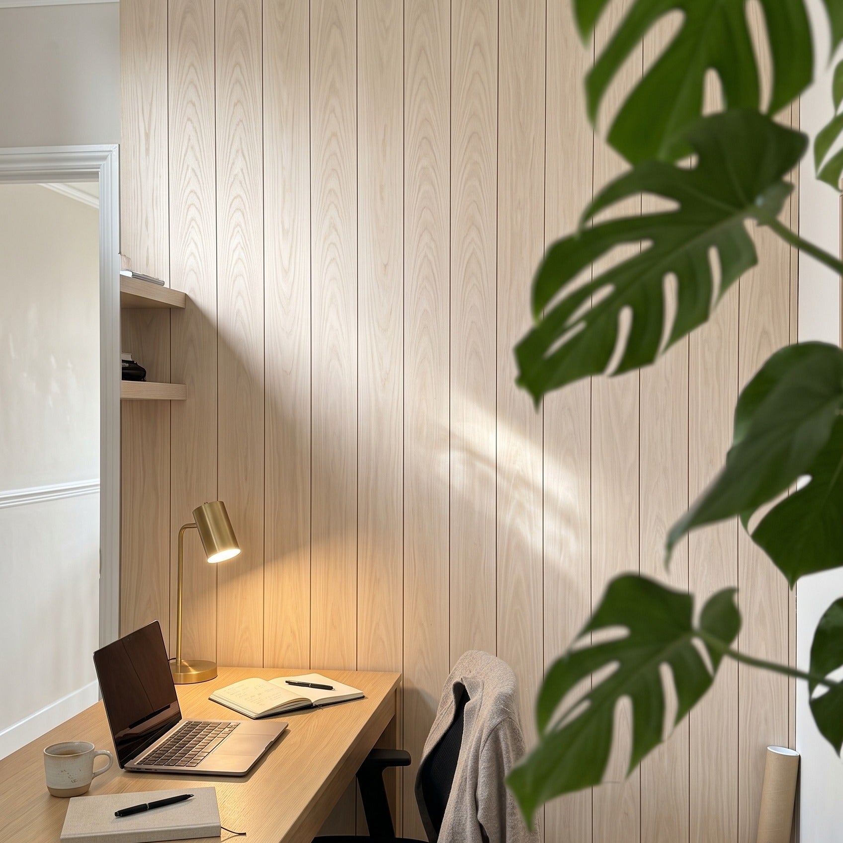 A cozy home office corner with a light wood desk, black office chair, brass desk lamp, laptop, and books against a light wood paneled wall, with a large green monstera leaf in the foreground.
